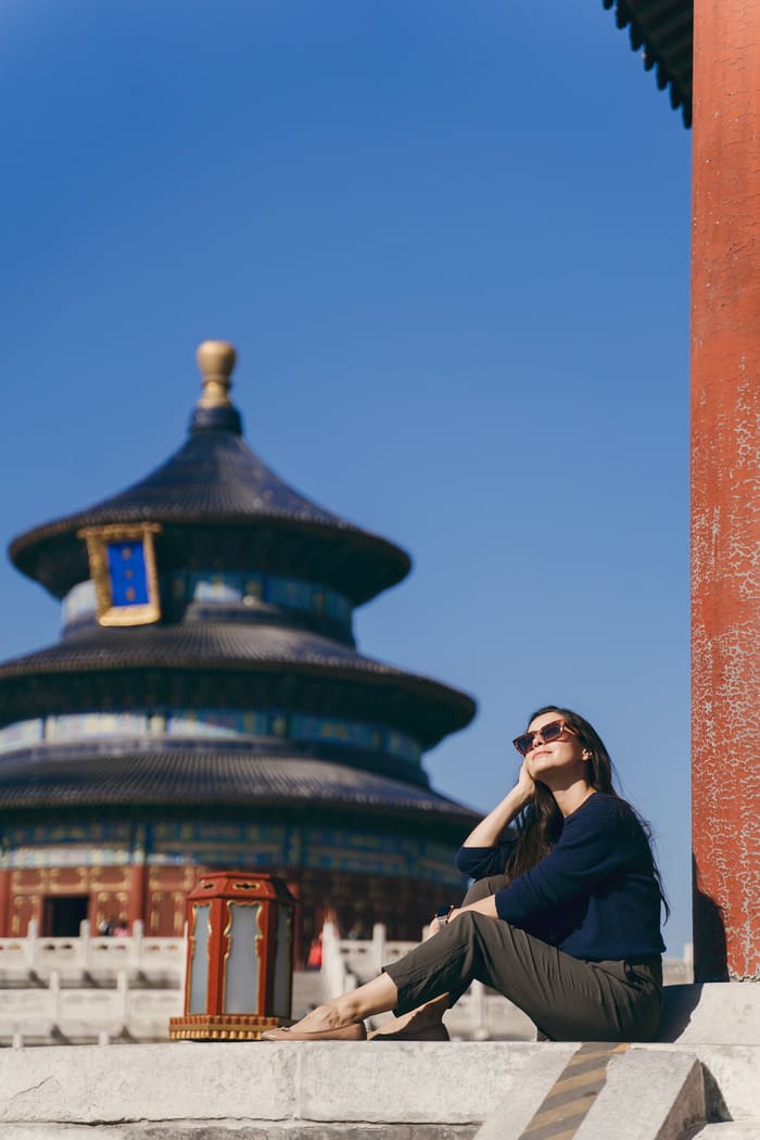 Una mujer con gafas de sol está sentada de perfil disfrutando del sol, con el Templo del Cielo de Pekín desenfocado al fondo bajo un cielo azul despejado.
