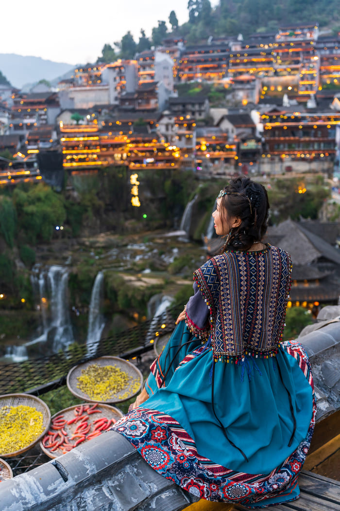 Una mujer con vestimenta tradicional de una minoría étnica china está sentada de perfil, contemplando un pueblo iluminado en la ladera de una montaña con cascadas al anochecer.