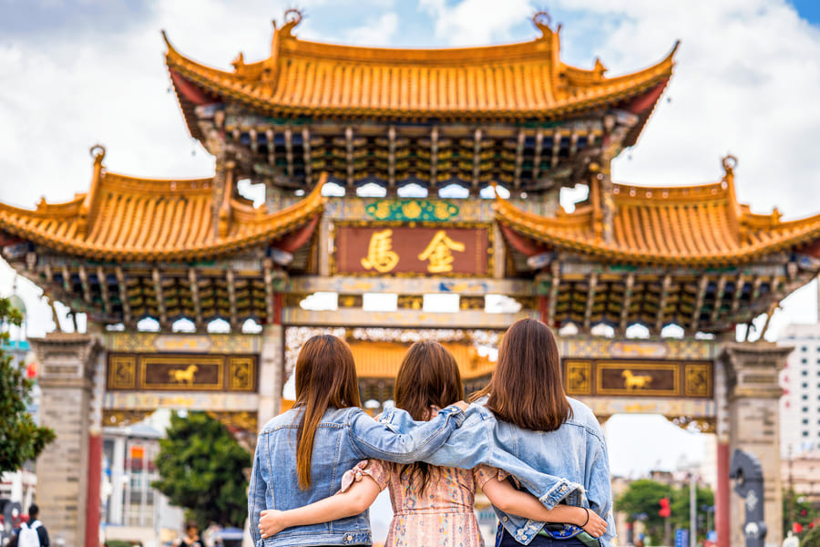 Tres mujeres de espaldas, abrazadas por los hombros y vistiendo chaquetas de mezclilla, contemplan una gran y ornamentada puerta ceremonial de arquitectura tradicional china con tejados dorados.