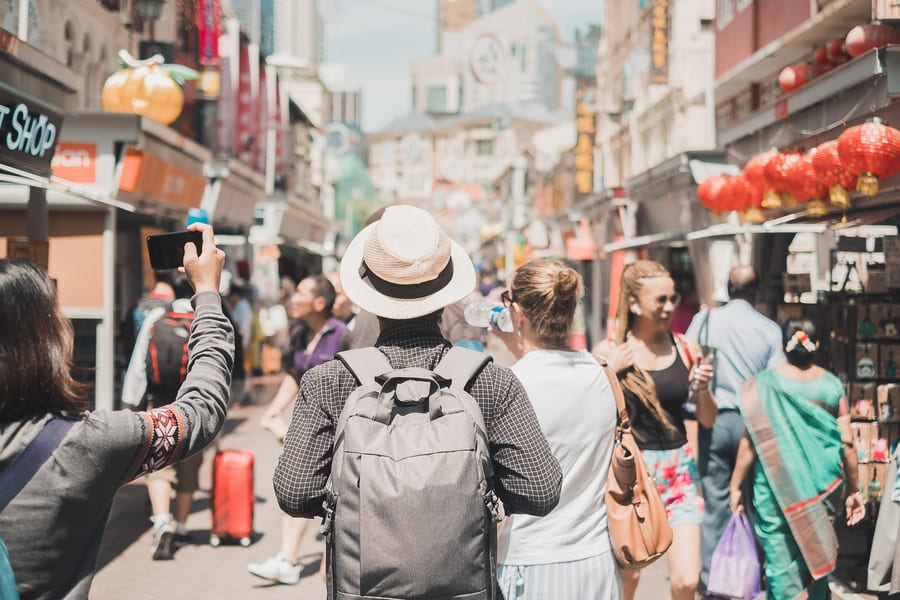 Vista desde atrás de un grupo de turistas caminando por una concurrida calle peatonal en un barrio asiático o Chinatown, decorada con farolillos rojos.